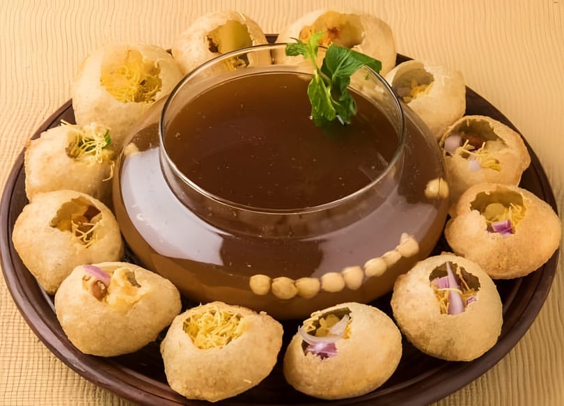 Crispy Pani Puri shells filled with savory ingredients, surrounding a bowl of mint-garnished tamarind water.